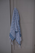 Blue DORA hand towel, 100x50 cm, displayed folded with soft fabric texture on a neutral background.
